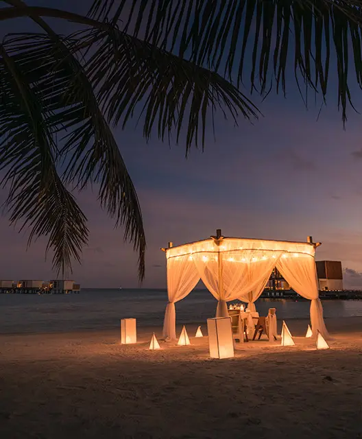 http://A%20romantic%20beach%20setup%20with%20a%20lit%20canopy%20and%20lanterns%20at%20sunset,%20surrounded%20by%20palm%20fronds%20and%20gentle%20waves%20in%20the%20background.