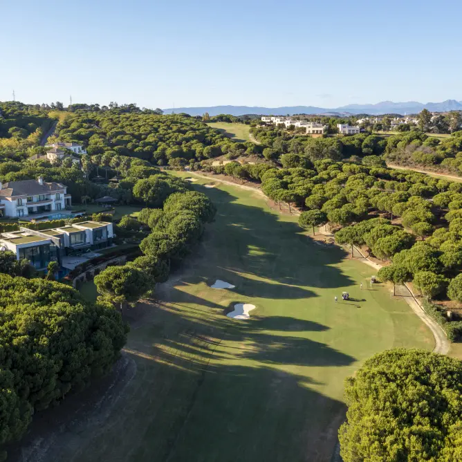 Aerial view of a lush golf course with verdant fairways and surrounding trees. To the left, a modern building overlooks the scenic landscape under a clear blue sky.