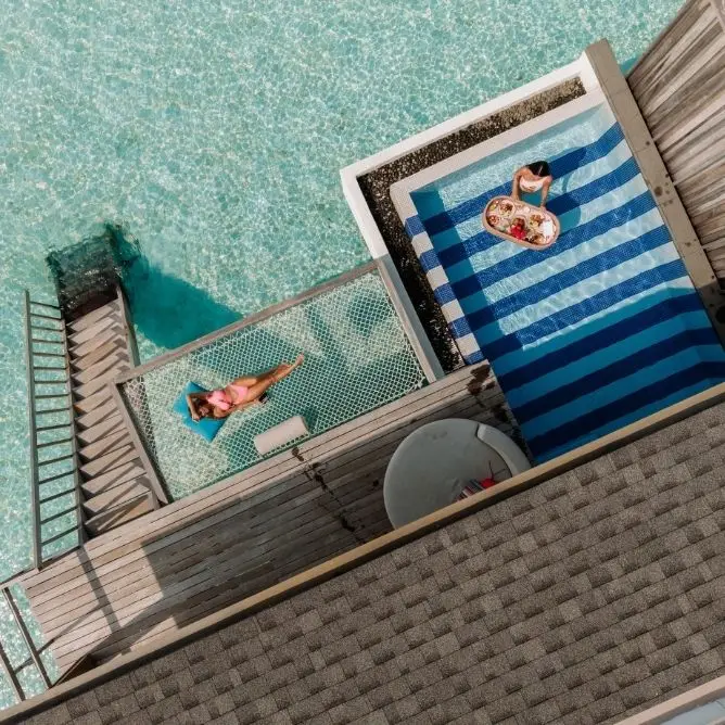 Overhead view of a woman relaxing on a net above clear water and another person with a floating tray of food in a pool next to a wooden deck.