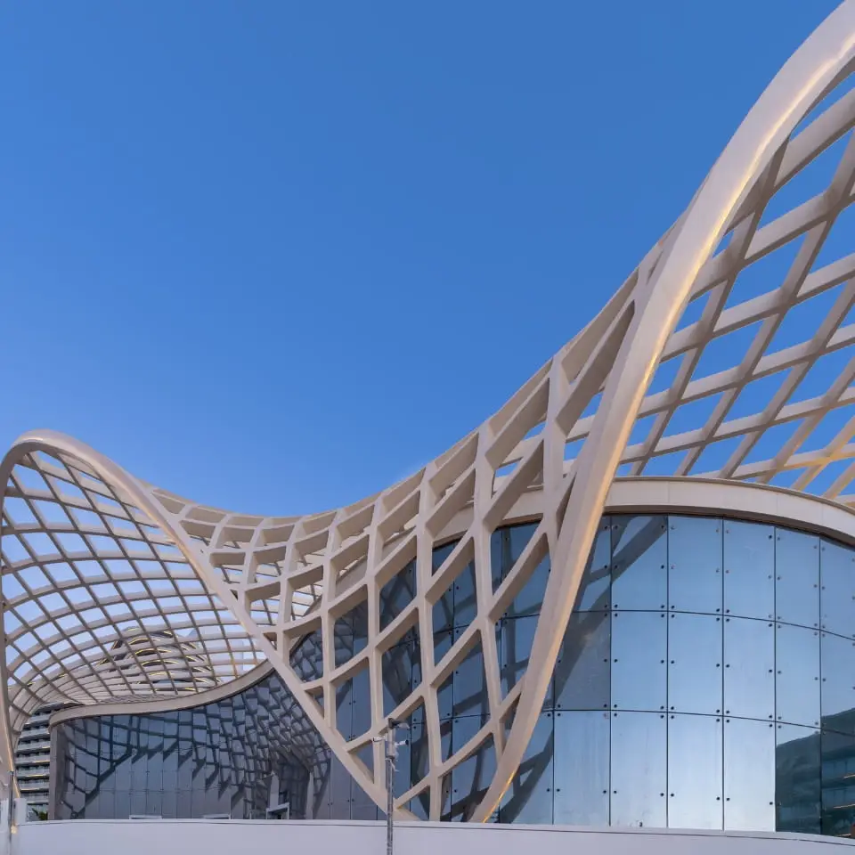 Modern architectural structure with a curved lattice roof above glass walls, set against a clear blue sky.