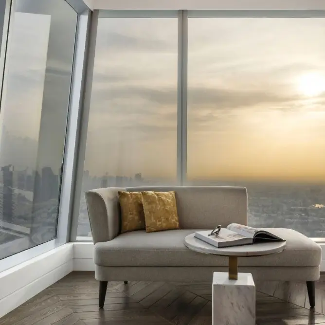A cozy gray couch with gold pillows sits near a small round table by a large window, overlooking a city skyline at sunset, creating a serene atmosphere.