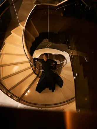 http://An%20elegant%20person%20in%20a%20long%20black%20dress%20lies%20gracefully%20on%20a%20spiral%20staircase,%20illuminated%20warmly,%20creating%20an%20artistic%20and%20contemplative%20atmosphere.