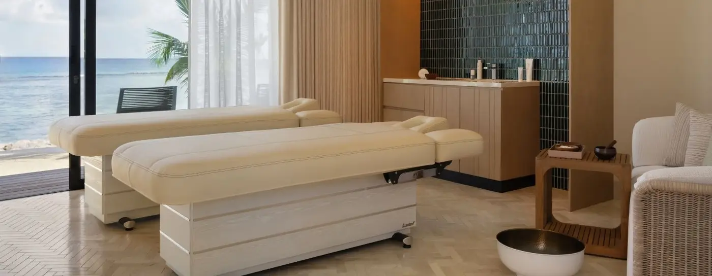 Bright spa room with two massage tables, large windows overlooking the ocean, and modern decor featuring natural light and greenery.