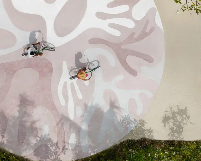 http://Two%20cyclists%20seen%20from%20above%20ride%20on%20a%20colorful,%20abstract-patterned%20surface,%20surrounded%20by%20shadows%20and%20greenery.