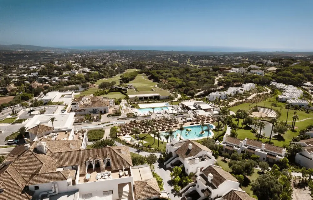 http://Aerial%20view%20of%20a%20luxurious%20resort%20with%20white%20buildings,%20two%20turquoise%20swimming%20pools,%20and%20lush%20greenery%20surrounded%20by%20a%20vast%20landscape%20under%20a%20clear%20blue%20sky.