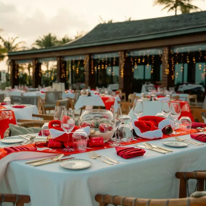 Outdoor resort restaurant tables set for a Christmas dinner with red runners, Santa hats, and festive centerpieces near the beach.