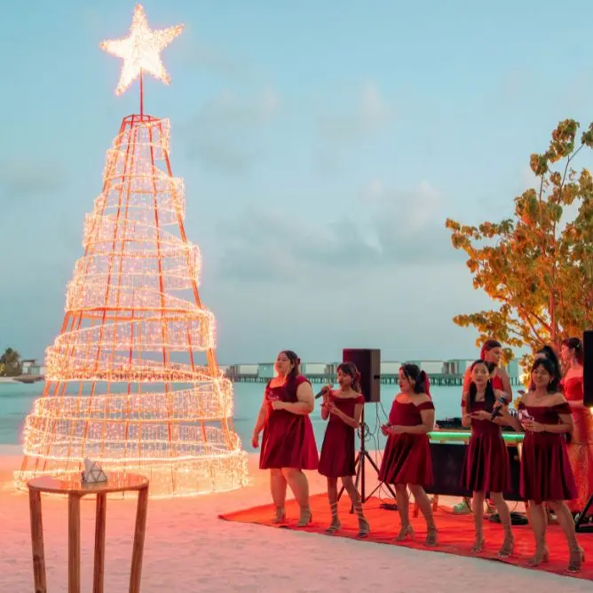 Group of women in red dresses singing next to a large spiral light Christmas tree on a sandy beach during a holiday celebration.
