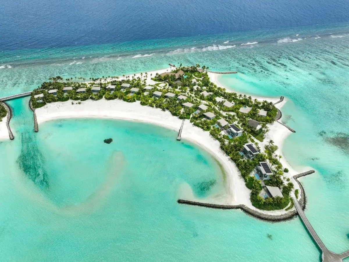 Aerial view of a tropical resort on a curved sandy peninsula, surrounded by turquoise waters and lush greenery.