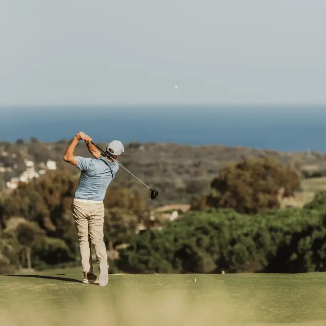 Golfer in light blue shirt and beige pants swinging club on green course with ocean and trees in the background.