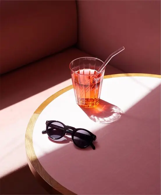 Une table élégante avec une surface rose sur laquelle sont posés un verre de boisson rouge avec une paille et des lunettes de soleil noires, projetant des ombres douces.
