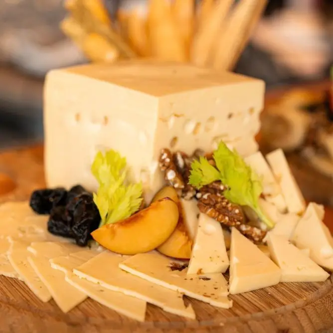 Gourmet cheese board with a large cheese block, sliced cheeses, dried plums, peaches, walnuts, and celery garnish.