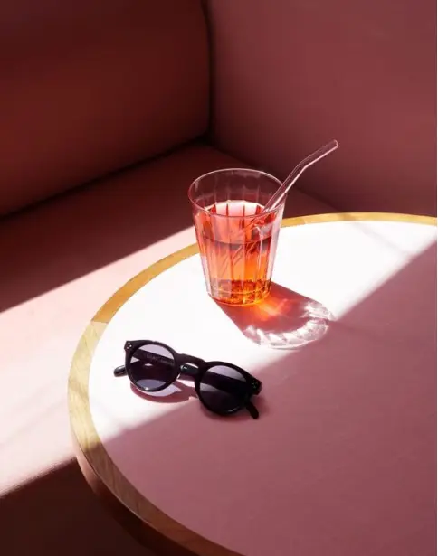 A stylish black pair of sunglasses rests on a pink table beside a glass of bright red drink with a straw, illuminated by sunlight.