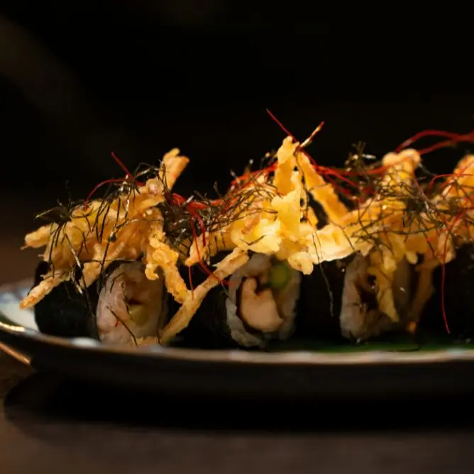 Modern fusion sushi rolls topped with crispy tempura shreds and delicate herb garnishes on a dark platter.