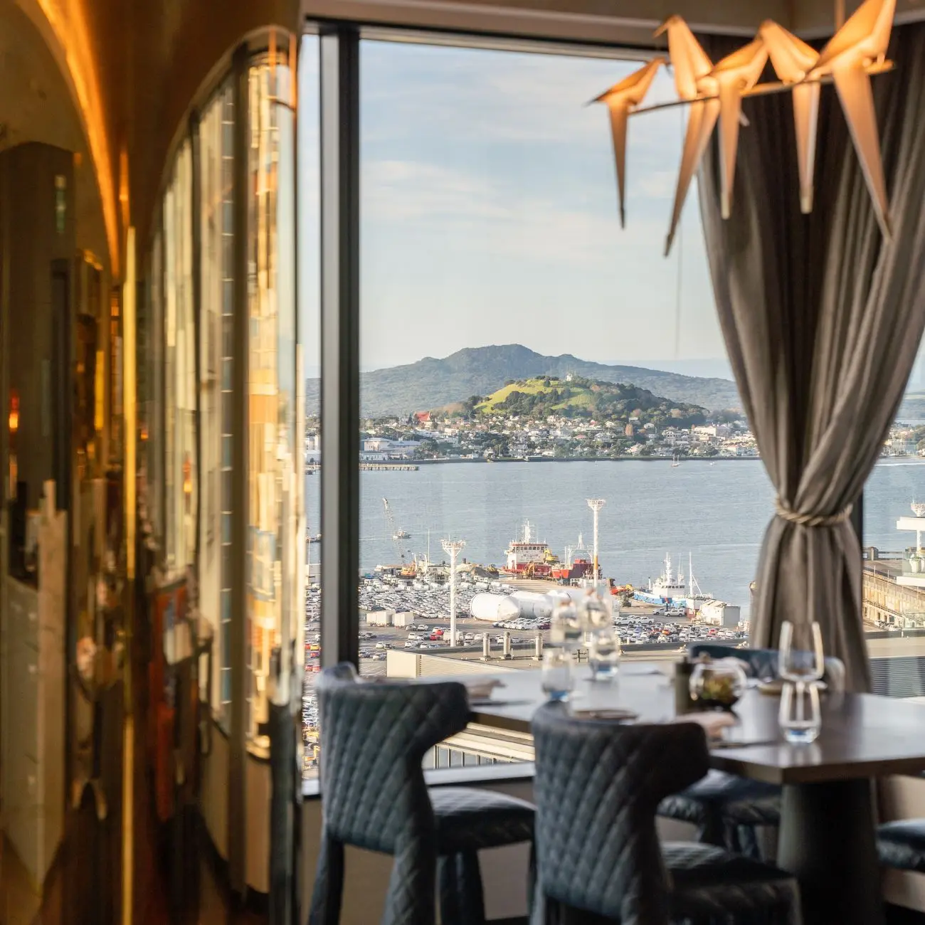 Elegant dining area with a view of the harbor, featuring boats and a mountain backdrop under a clear sky. Modern decor and ambiance.