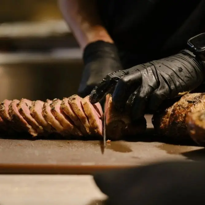 Close-up of a chef wearing black gloves slicing a tender piece of medium-rare roast meat on a cutting board.