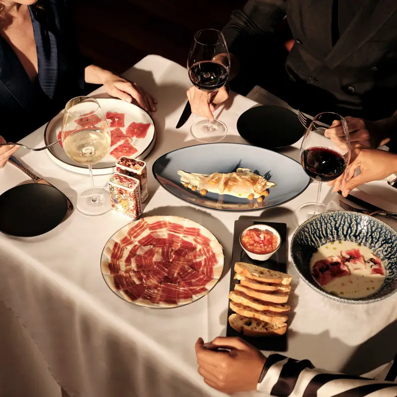 A candlelit dinner table with plates of charcuterie, a seafood dish, and breadsticks. Three people toast with red wine, creating a cozy, elegant atmosphere.