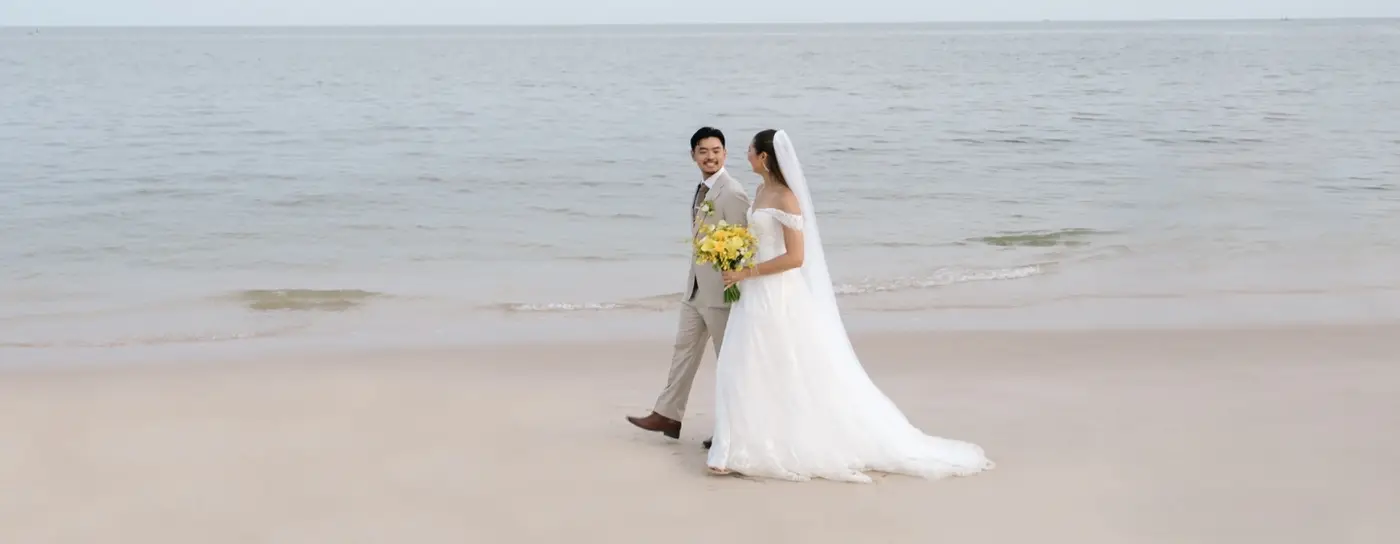 A bride in a flowing white gown holds yellow flowers, walking hand-in-hand with the groom in a light suit along a serene beach by the ocean. Romantic ambiance.