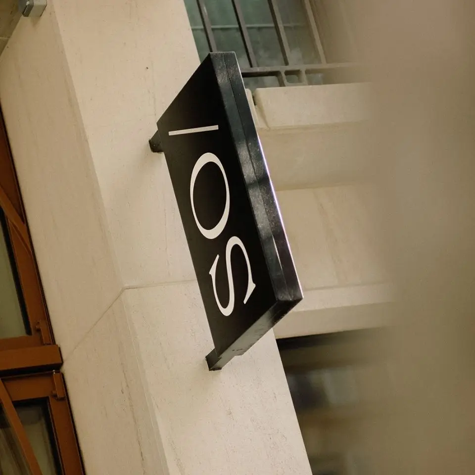 Black rectangular sign with white text displaying "SO/" mounted on a beige building wall near windows.