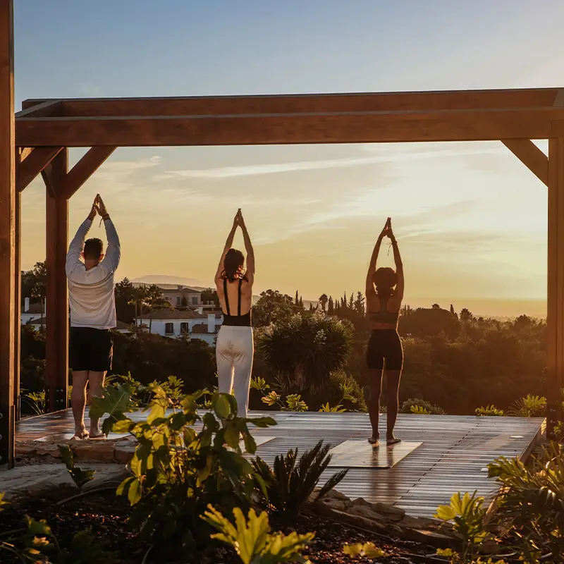 Three people practice yoga at sunset under a wooden pergola, with lush greenery and distant hills in the background, conveying tranquility and focus.