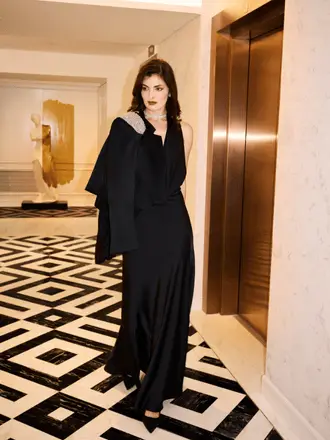 http://A%20woman%20in%20a%20long%20black%20dress%20stands%20confidently%20in%20a%20stylish%20hotel%20lobby.%20She%20has%20a%20jacket%20draped%20over%20her%20shoulder.%20The%20floor%20features%20a%20geometric%20black-and-white%20pattern,%20and%20an%20elevator%20with%20closed%20gold%20doors%20is%20in%20the%20background.