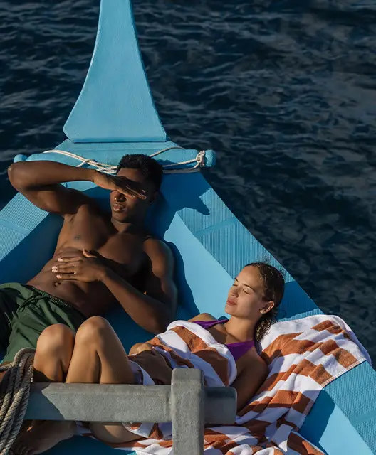 http://A%20couple%20relaxes%20on%20a%20blue%20boat,%20lying%20on%20striped%20towels,%20surrounded%20by%20calm%20ocean%20waters%20under%20sunlight.