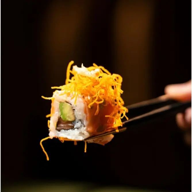 Close-up of a salmon avocado sushi roll garnished with bright orange crispy tempura flakes held by chopsticks.