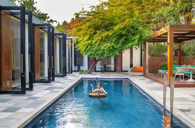 http://A%20person%20relaxes%20on%20a%20float%20with%20food%20in%20a%20modern%20pool%20surrounded%20by%20open%20patio%20doors.%20A%20lush%20tree%20and%20outdoor%20seating%20create%20a%20tranquil%20vibe.