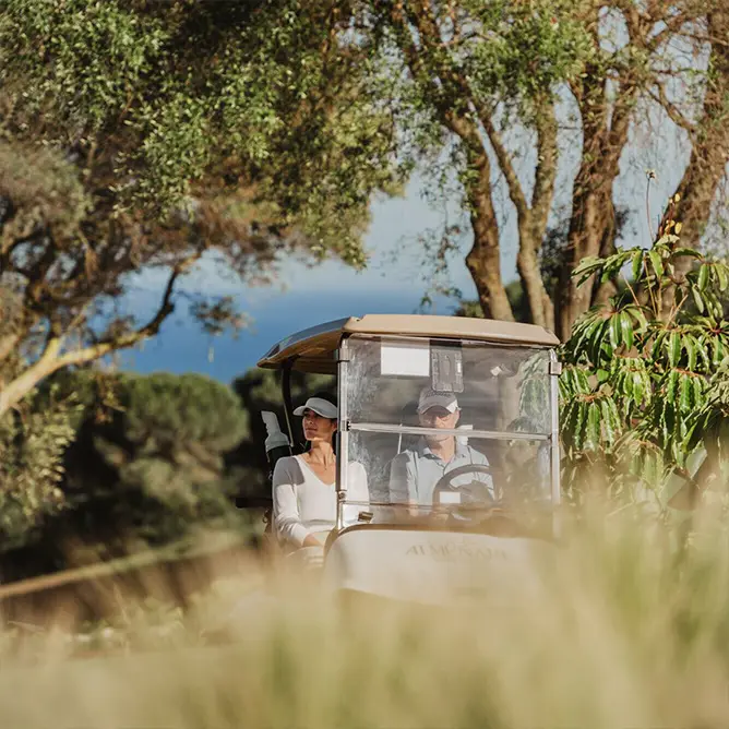 A golf cart travels on a sunlit path surrounded by trees and tall grass, creating a relaxed and serene outdoor atmosphere.