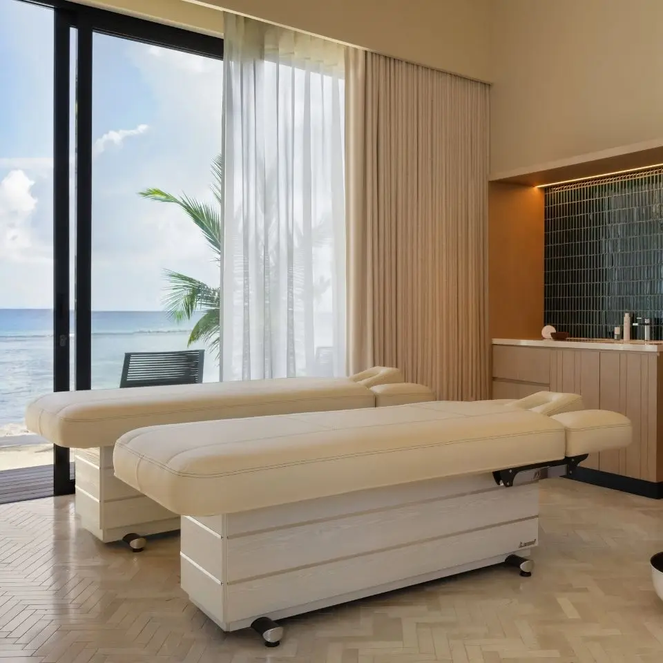 Bright spa room with two massage tables, large windows overlooking the ocean, and modern decor featuring natural light and greenery.