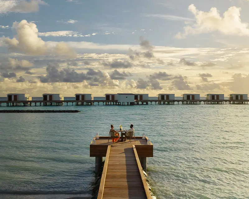 http://Two%20people%20sit%20at%20the%20end%20of%20a%20wooden%20pier%20overlooking%20the%20ocean,%20with%20overwater%20bungalows%20in%20the%20background%20under%20a%20colorful%20sunset%20sky.