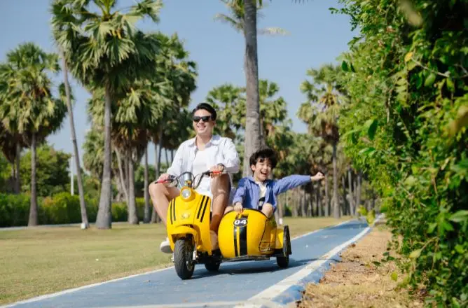 http://A%20smiling%20father%20and%20his%20young%20son%20riding%20a%20fun%20yellow%20scooter%20with%20a%20sidecar%20along%20a%20blue%20paved%20path%20lined%20with%20tall%20palm%20trees.