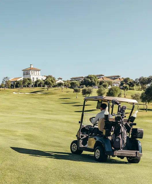 A man driving a golf cart across a lush green golf course under a clear blue sky.
