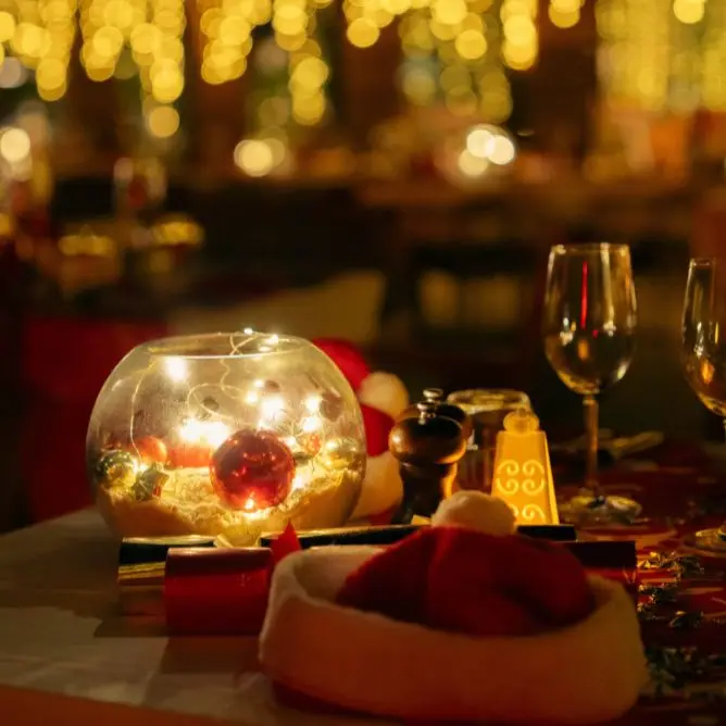Festive Christmas dining table setup featuring a glowing glass centerpiece filled with ornaments, a Santa hat, and wine glasses against a bokeh background of holiday lights.