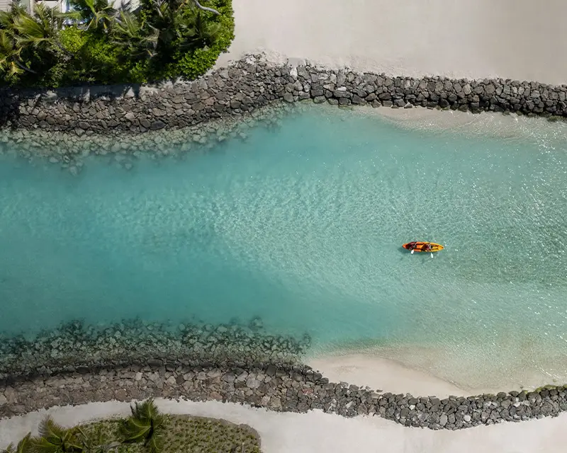 http://Aerial%20view%20of%20a%20tranquil%20turquoise%20waterway%20bordered%20by%20gray%20stones%20and%20lush%20greenery,%20with%20a%20lone%20kayak%20in%20the%20center.